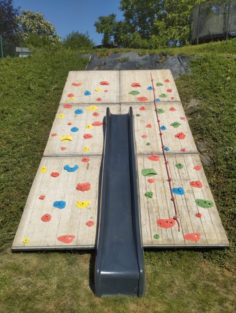 Vooraanzicht van de afgewerkte klimhelling in de tuin met glijbaan in het midden en kleurrijke grepen