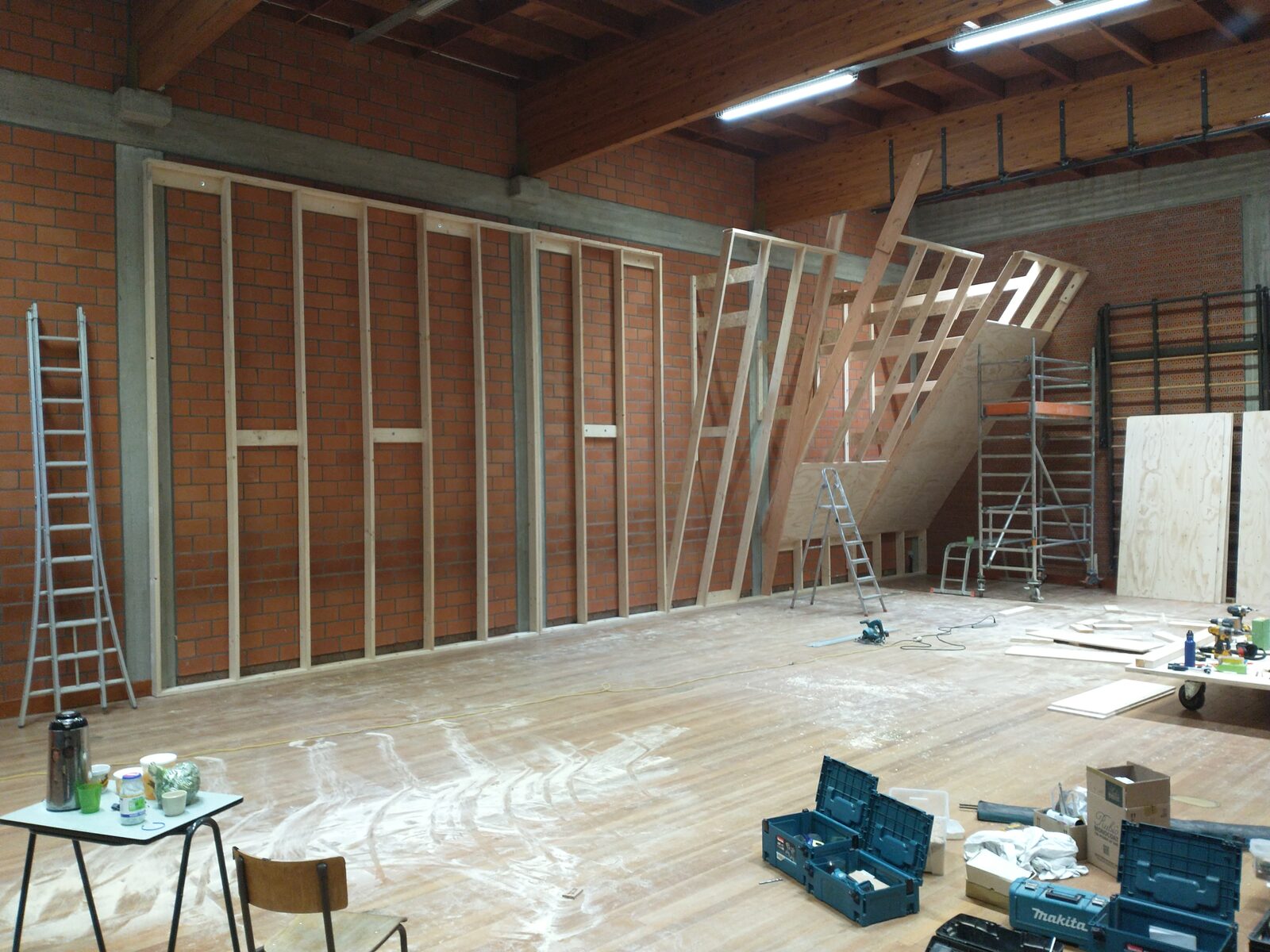 Houten draagstructuur van een klimmuur in opbouw tegen een bakstenen muur, met rolsteiger, ladders en multiplexplaten
